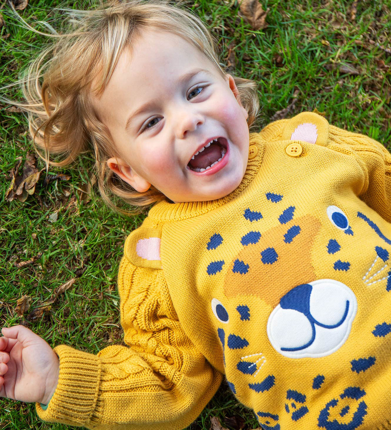 A child laying on grass wearing the Frugi Caleb Leopard Character Knit Jumper. This knitted jumper is a part of a wide range of organic cotton clothing for children available here at Babipur. 