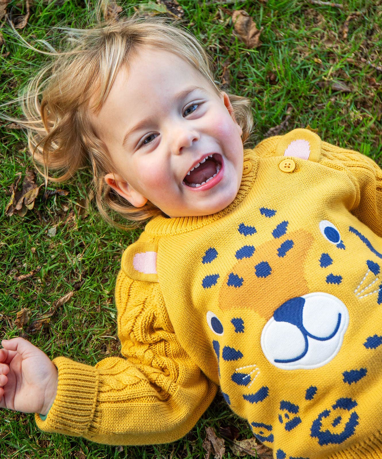 A child laying on grass wearing the Frugi Caleb Leopard Character Knit Jumper. This knitted jumper is a part of a wide range of organic cotton clothing for children available here at Babipur. 