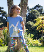 Child leaning against a tree outside wearing the Frugi Cassia t-shirt.  Cornflower blue coloured with a playful appliqué of a grey rabbit with flowers in its mouth with the Frugi Libby Leggings with a floral themed Bunny Bounce print. 