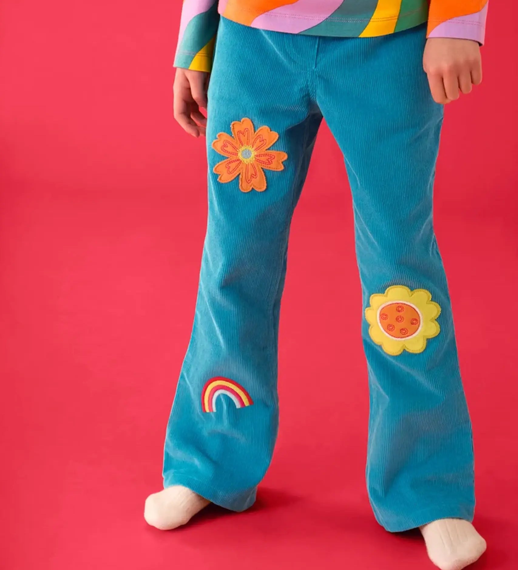 Close up of a child wearing the Frugi cherry boat blue corduroy flared trousers  with two flowers and a rainbow badge style details.