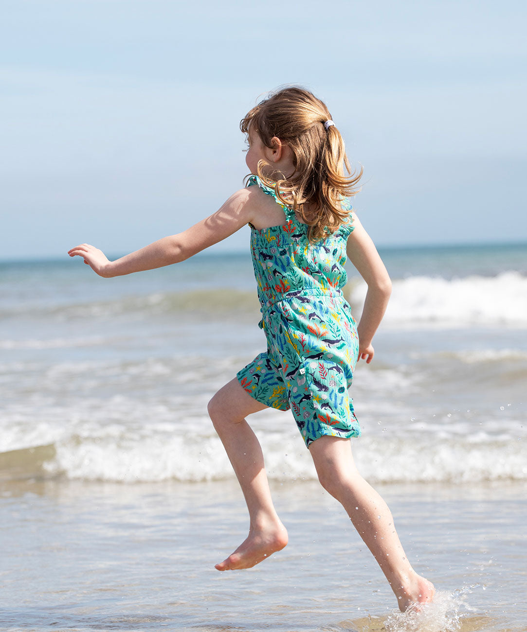 A child facing away fro,m teh camera splashing around in teh sea wearing a Frugi Children's Maris Playsuit with a  Sea Life print.