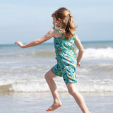 A child facing away fro,m teh camera splashing around in teh sea wearing a Frugi Children's Maris Playsuit with a  Sea Life print.