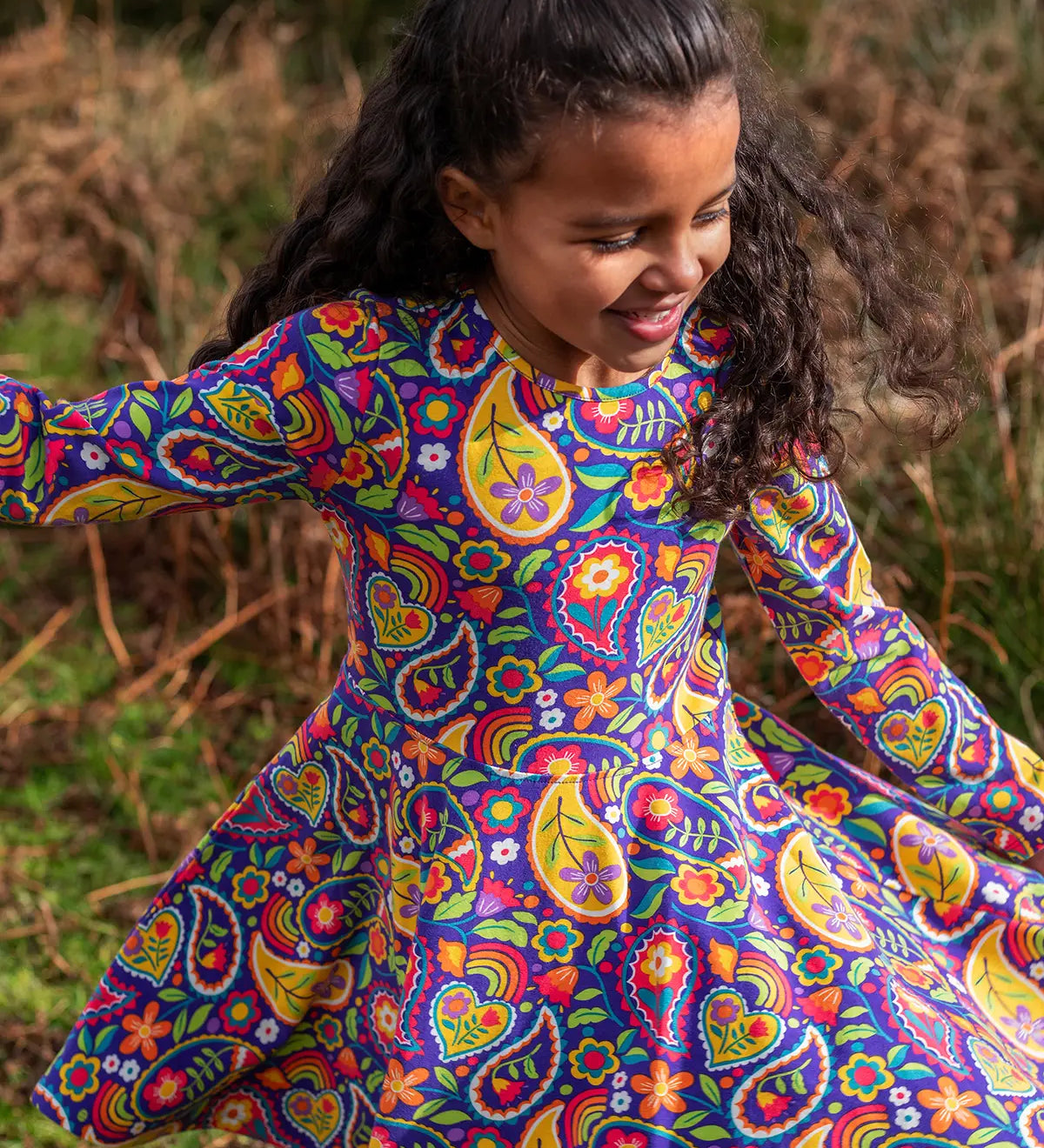 Child wearing the Frugi GOTS organic cotton sofia skater dress in pretty paisley. Purple background with colourful paisley print featuring flower and rainbow details.