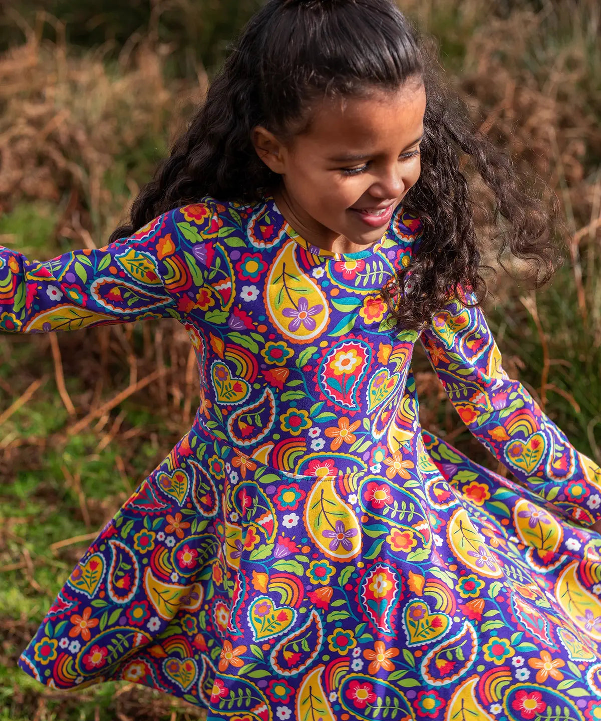 Child wearing the Frugi GOTS organic cotton sofia skater dress in pretty paisley. Purple background with colourful paisley print featuring flower and rainbow details.