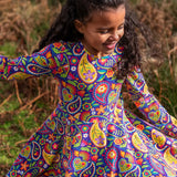 Child wearing the Frugi GOTS organic cotton sofia skater dress in pretty paisley. Purple background with colourful paisley print featuring flower and rainbow details.