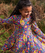 Child wearing the Frugi GOTS organic cotton sofia skater dress in pretty paisley. Purple background with colourful paisley print featuring flower and rainbow details.