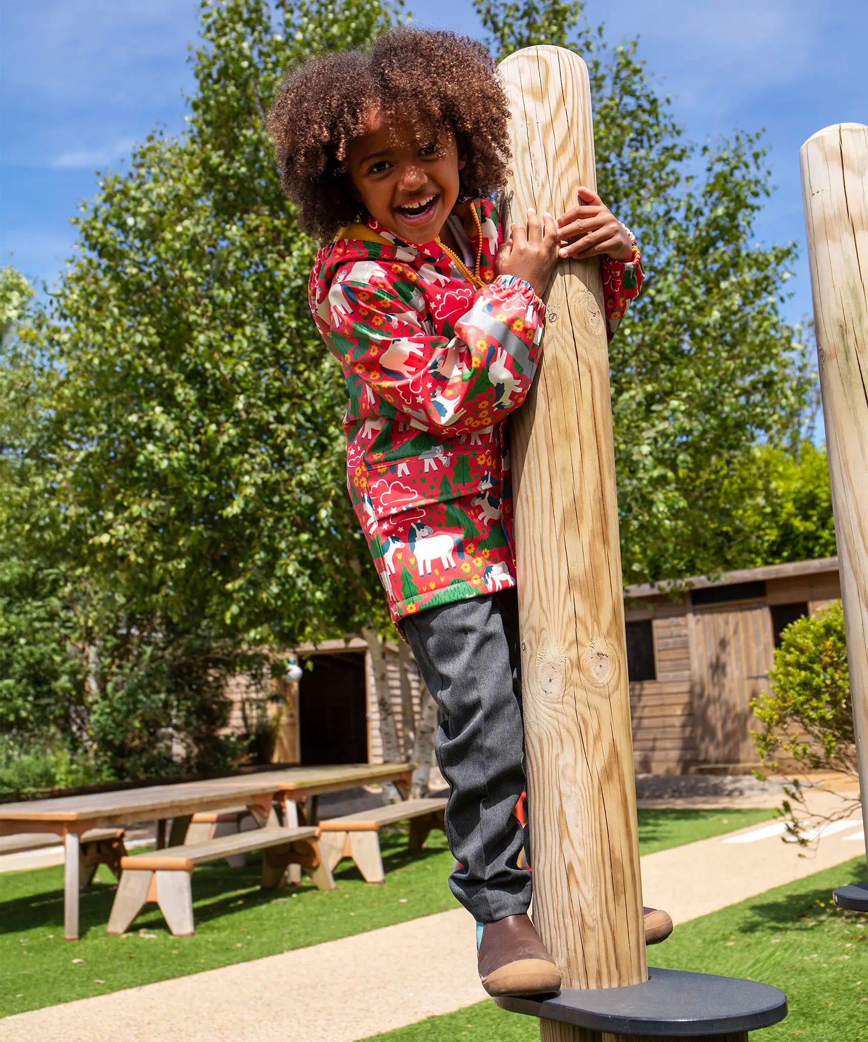 Child wearing Frugi waterproof puddle buster coat in pink purrfect ponies print, standing on a platform in a playground.