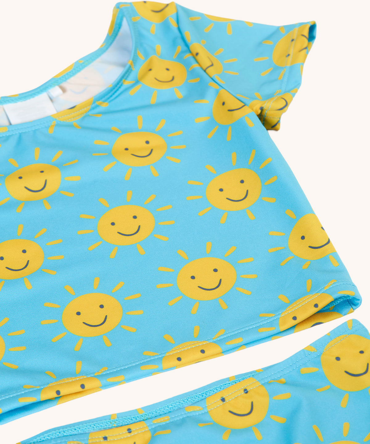 A close up of the Frugi blue children's tankini with an all over yellow smiley sunshine print. A short sleeved swimwear top with matching brief style bottoms.