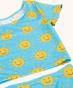 A close up of the Frugi blue children's tankini with an all over yellow smiley sunshine print. A short sleeved swimwear top with matching brief style bottoms.