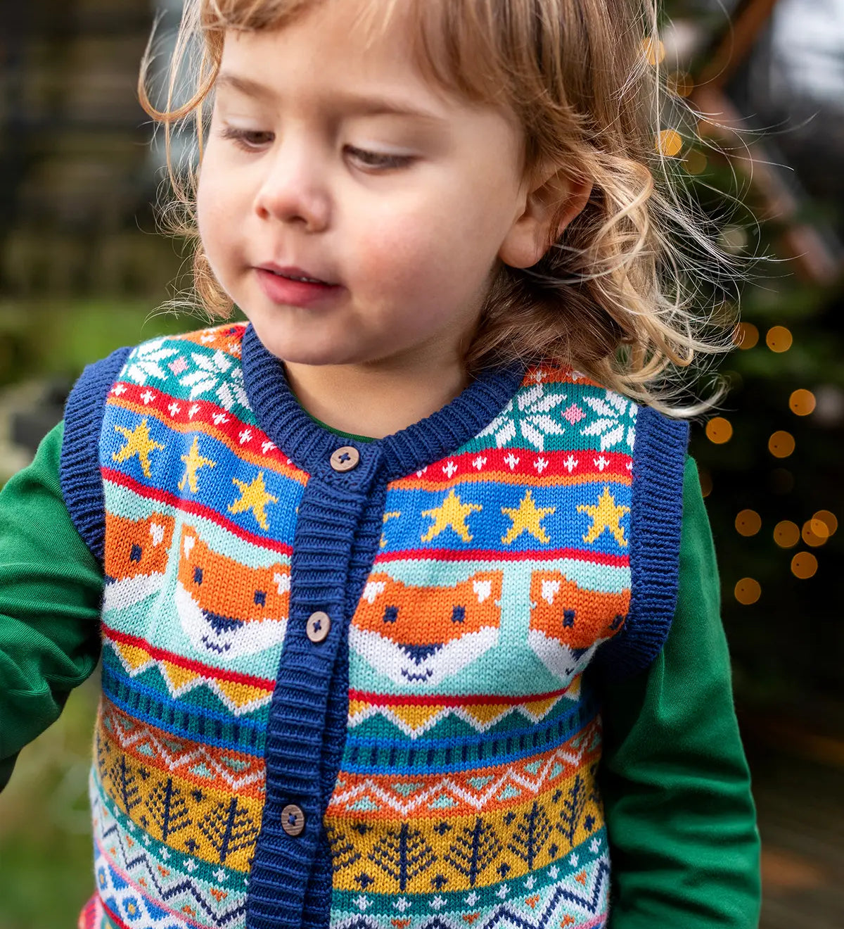 Child wearing the Frugi fox fair isle organic cotton wenn knitted waistcoat. Multi-coloured fair isle knit with navy edges and wooden buttons with a long sleeved green top underneath.