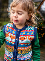 Child wearing the Frugi fox fair isle organic cotton wenn knitted waistcoat. Multi-coloured fair isle knit with navy edges and wooden buttons with a long sleeved green top underneath.