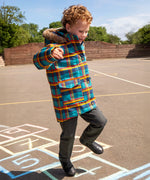 Child wearing Frugi's Penhale parka coat in a blue red and yellow tartan check with the faux fur trimmed hood down playing hopscotch in a playground.