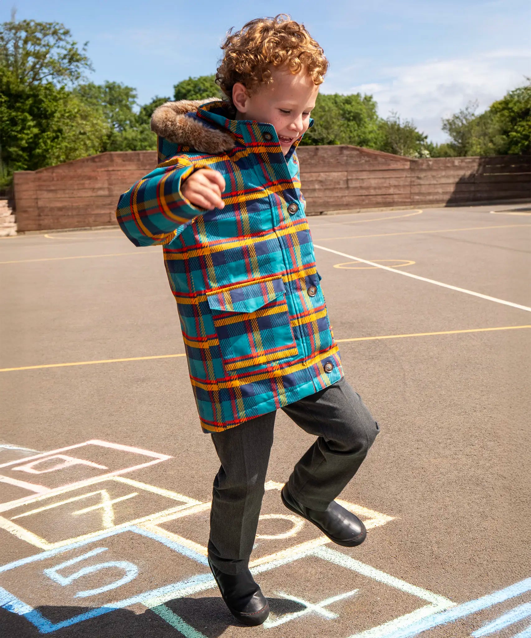 Child wearing Frugi's Penhale parka coat in a blue red and yellow tartan check with the faux fur trimmed hood down playing hopscotch in a playground.