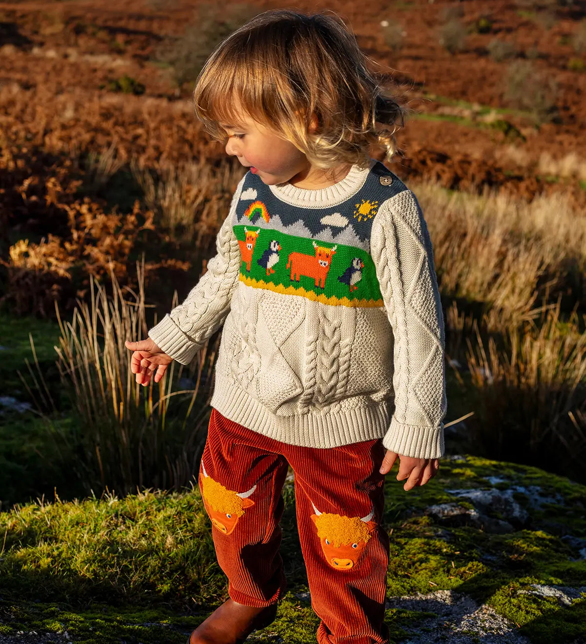 Child wearing Frugi clyde cable knit organic cotton jumper natural highlands cow and puffin design with rust highland cow corduroy trousers.