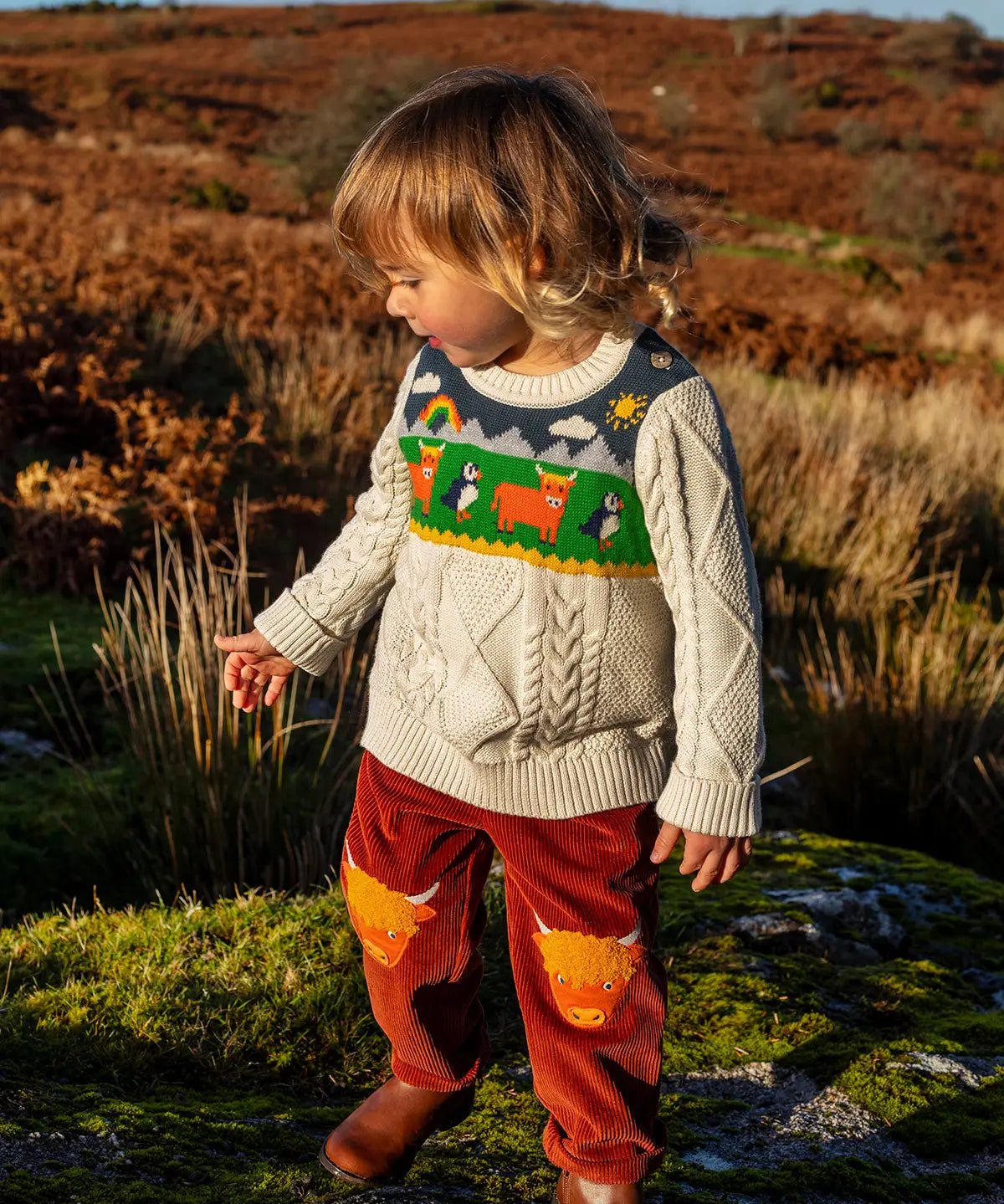 Child wearing Frugi clyde cable knit organic cotton jumper natural highlands cow and puffin design with rust highland cow corduroy trousers.