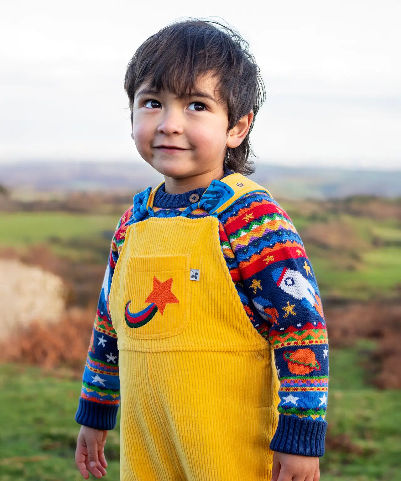 Child wearing the Frugi Gylly cord reversible dungaree in bumblebee yellow with star pocket design with a fair isle knitted jumper underneath. 