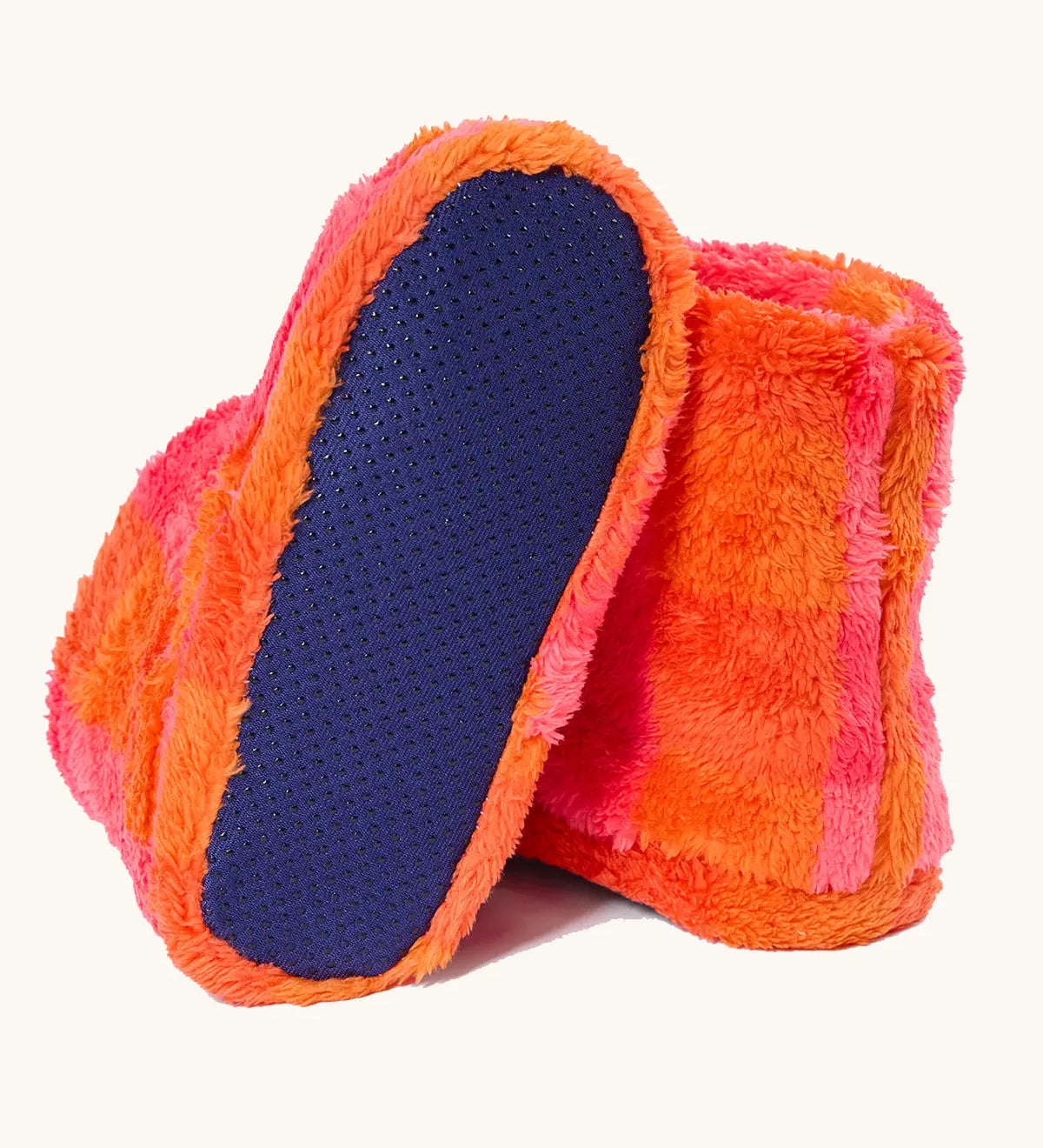 A pair of frugi fuzzy slipper boots in a bright orange and pink striped pattern, with one boot turned to show the dark blue textured non-slip sole.