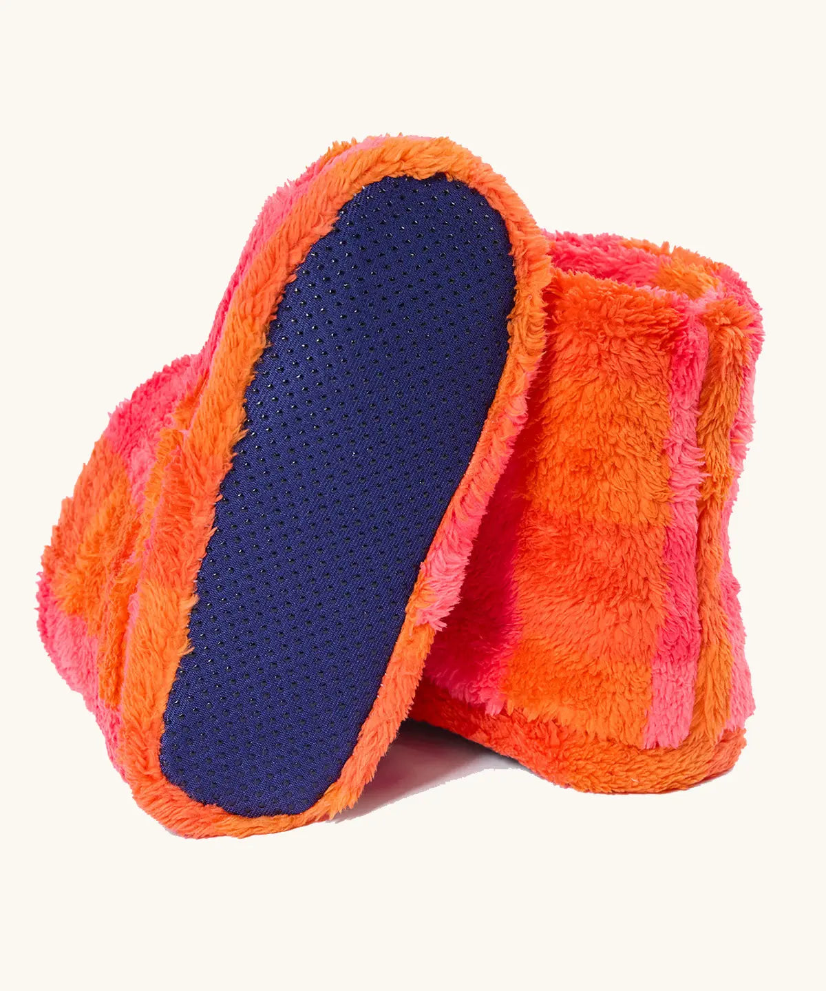 A pair of frugi fuzzy slipper boots in a bright orange and pink striped pattern, with one boot turned to show the dark blue textured non-slip sole.