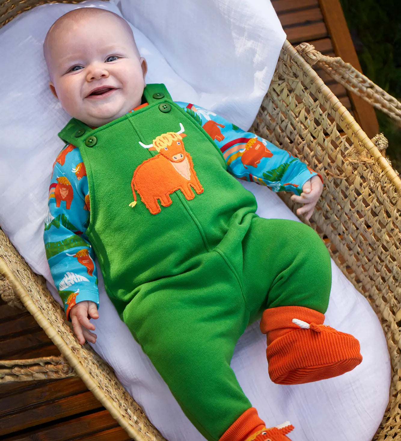 Baby wearing the Frugi coverack palm green dungaree with highland coos applique with a highland cows design long sleeve body underneath. 