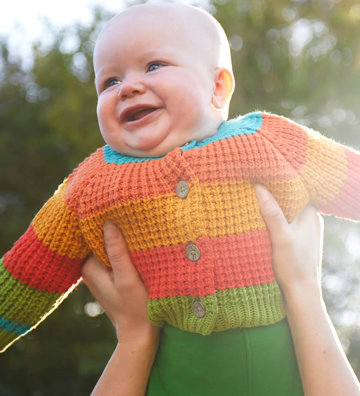 Baby wearing the Frugi cuddly knit highland wide stripe rainbow cardigan being held up. 