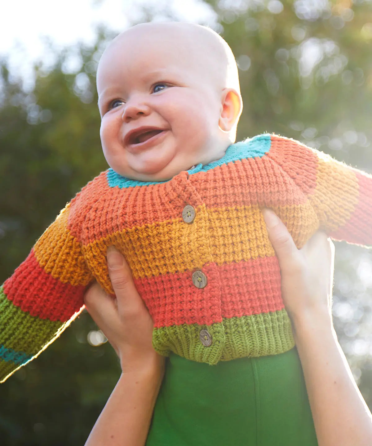Baby wearing the Frugi cuddly knit highland wide stripe rainbow cardigan being held up. 