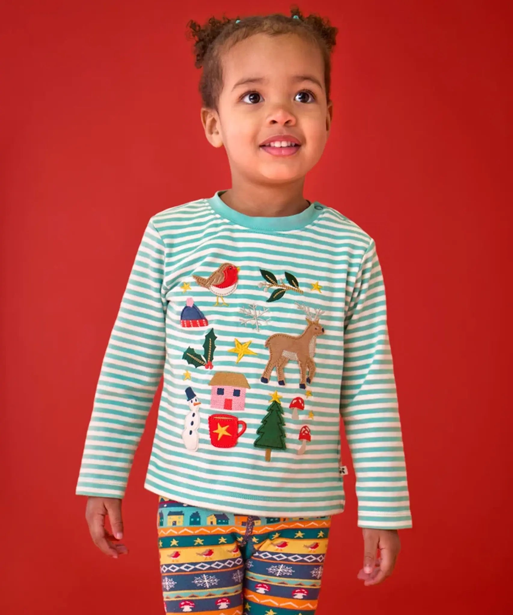 A young child wearing a Frugi organic cotton long-sleeve striped top with a festive Christmas-themed applique design and knitted fair isle leggings.