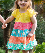 A smiling toddler wearing Frugi Dorothy Twirly Dress with a Rainbow Hotchpotch print, a patchwork type design featuring yellow, red orange and green patterns with daisies, running amongst trees.