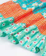 A close up of Frugi Dorothy Twirly Dress, with a Rainbow Hotchpotch print, a patchwork type design featuring orange, mint and green patterns with daisies.