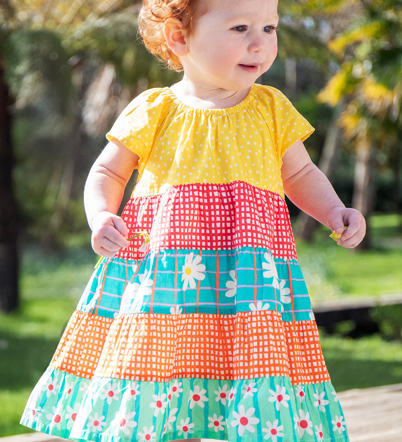 A toddler wearing the Frugi Dorothy Twirly Dress, with a Rainbow Hotchpotch print, a patchwork type design featuring yellow, red orange and green patterns with daisies, stood amongst trees.