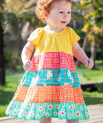 A toddler wearing the Frugi Dorothy Twirly Dress, with a Rainbow Hotchpotch print, a patchwork type design featuring yellow, red orange and green patterns with daisies, stood amongst trees.