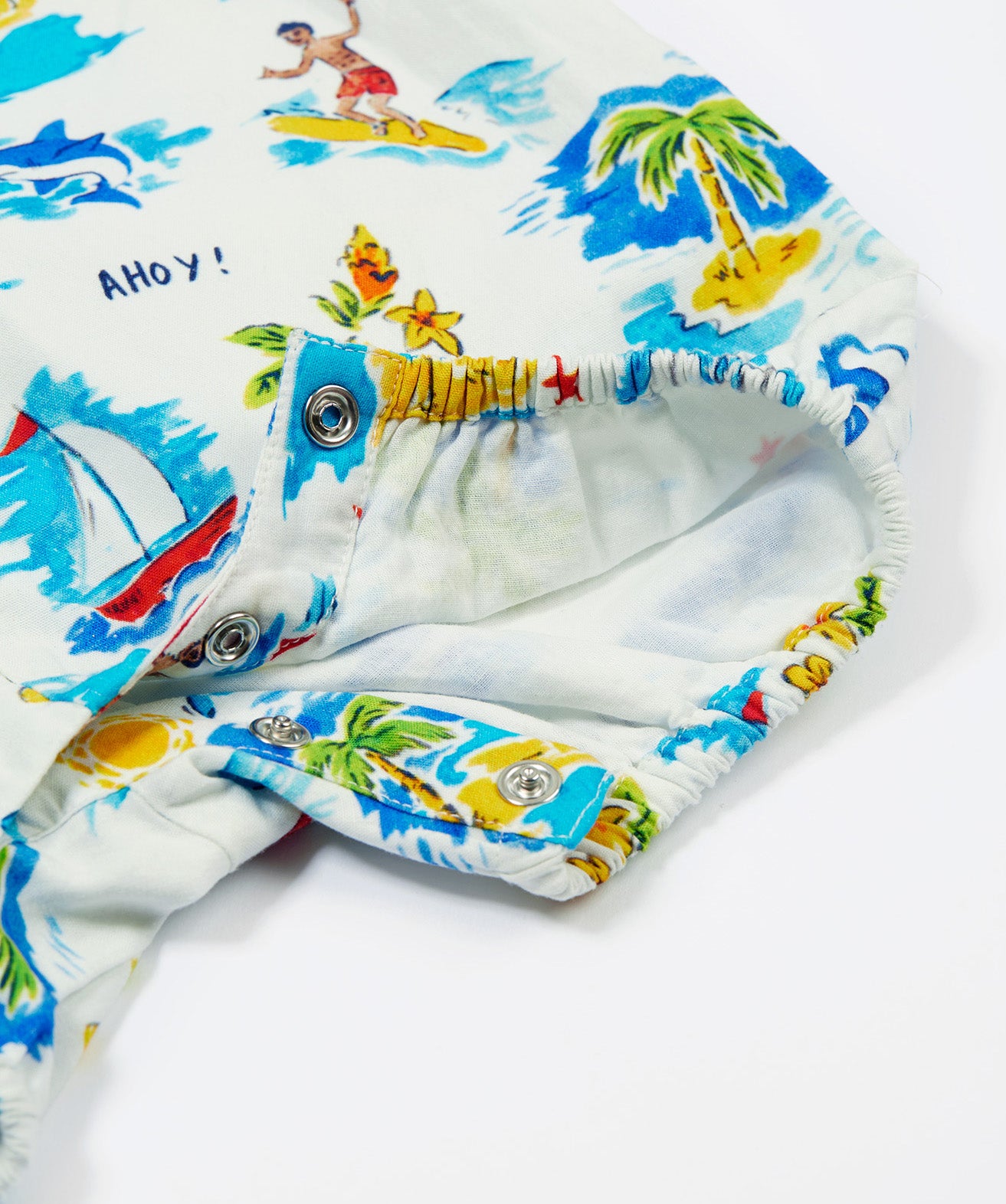 A close-up of the Frugi Aloha Organic Cotton Dungarees popper fastenings, featuring a colourful tropical beach print with dolphins, rainbows and sailing boats.