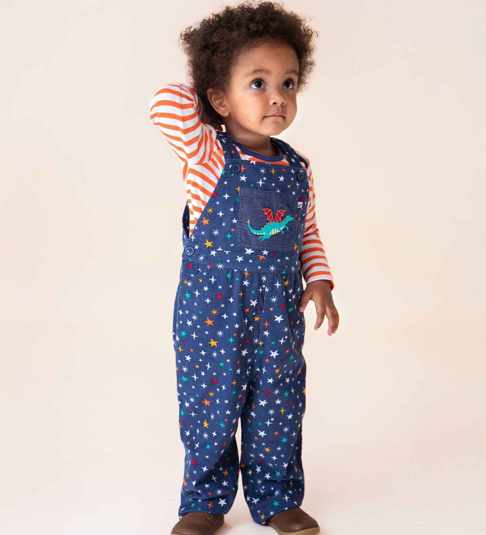 A child wearing the Frugi  Rainbow Galaxy Dragon Durgan Cotton Corduroy Dungarees with a white and orange striped top underneath. These navy cord dungarees have a rainbow coloured star print all over and a green dragon applique on the front pocket. These dungarees are part of a wide range of organic cotton clothing for children available here at Babipur