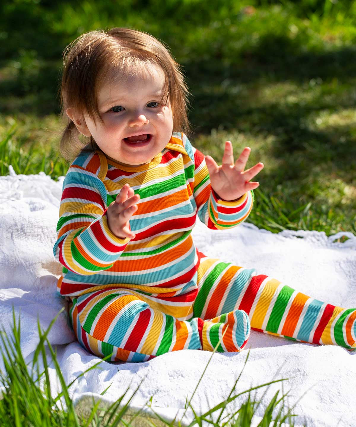 A baby sitting up on a blanket placed on grass outdoors wearing a Frugi easy dressing rainbow striped ribbed material babygrow. 