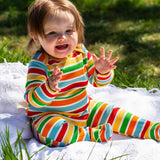 A baby sitting up on a blanket placed on grass outdoors wearing a Frugi easy dressing rainbow striped ribbed material babygrow. 