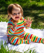 A baby sitting up on a blanket placed on grass outdoors wearing a Frugi easy dressing rainbow striped ribbed material babygrow. 