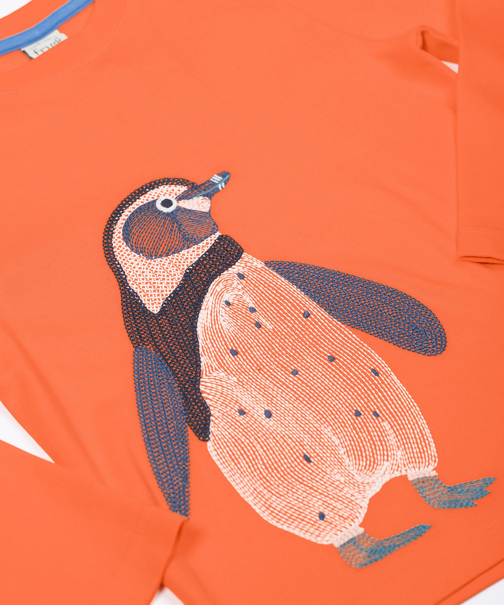 A close up of the Frugi kids embroidered adventure top in salamander/penguin showing the full penguin embroidery detail on pastel orange organic cotton fabric.