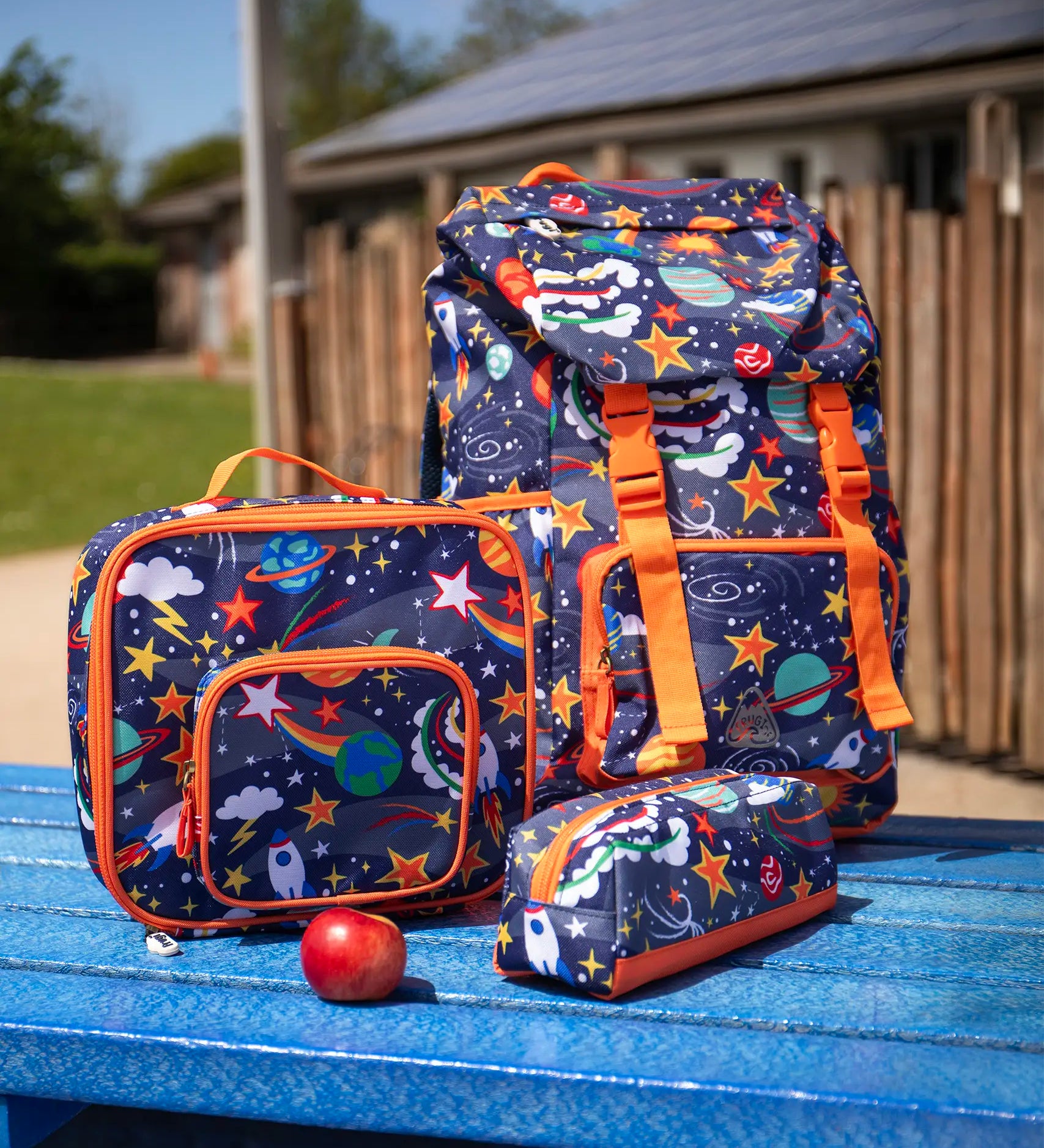 Frugi trail blazing backpack, lunch bag and matching pencil case in the explore the stars design placed on a blue picnic table.