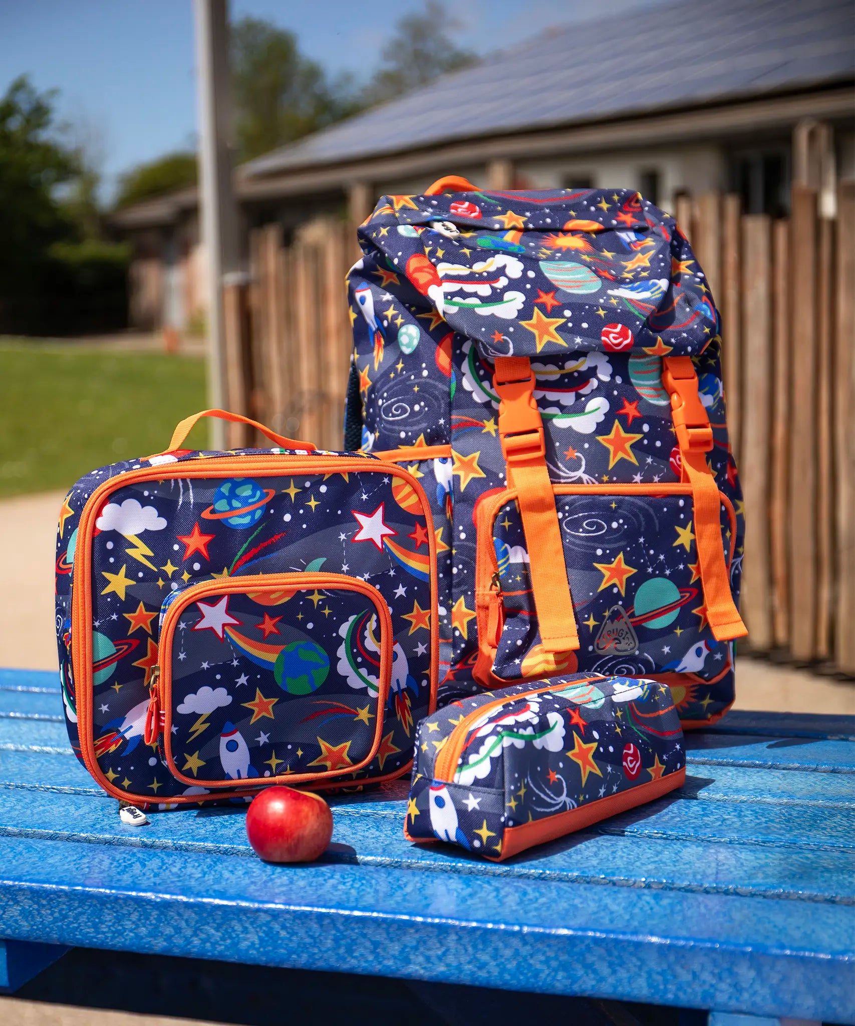 Frugi trail blazing backpack, lunch bag and matching pencil case in the explore the stars design placed on a blue picnic table.