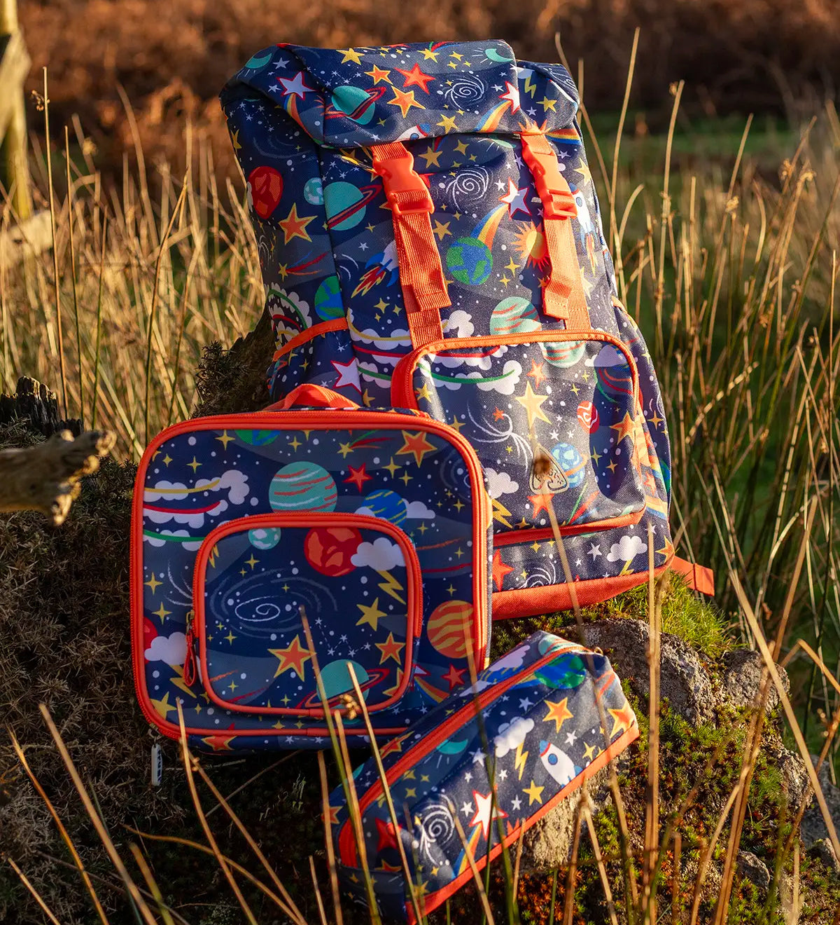 Frugi trail blazing backpack, lunch bag and pencil case in the explore the stars design standing on a rock in a grassy field