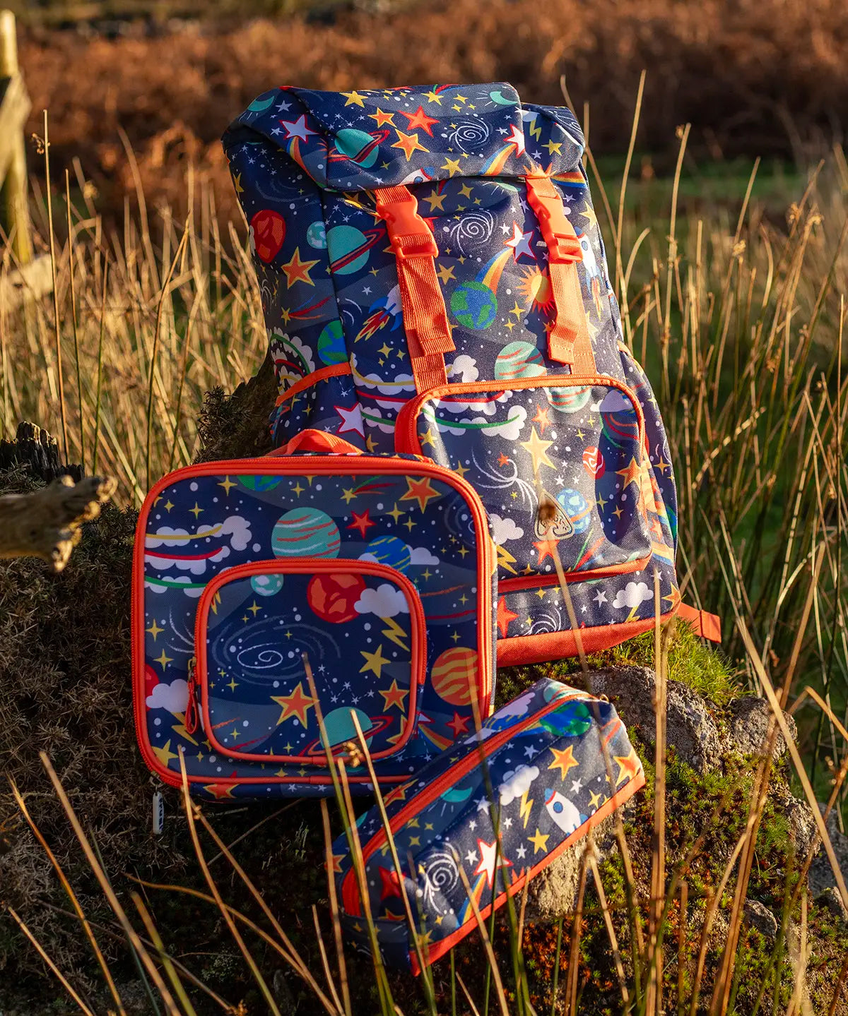 Frugi trail blazing backpack, lunch bag and pencil case in the explore the stars design standing on a rock in a grassy field