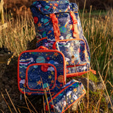Frugi trail blazing backpack, lunch bag and pencil case in the explore the stars design standing on a rock in a grassy field