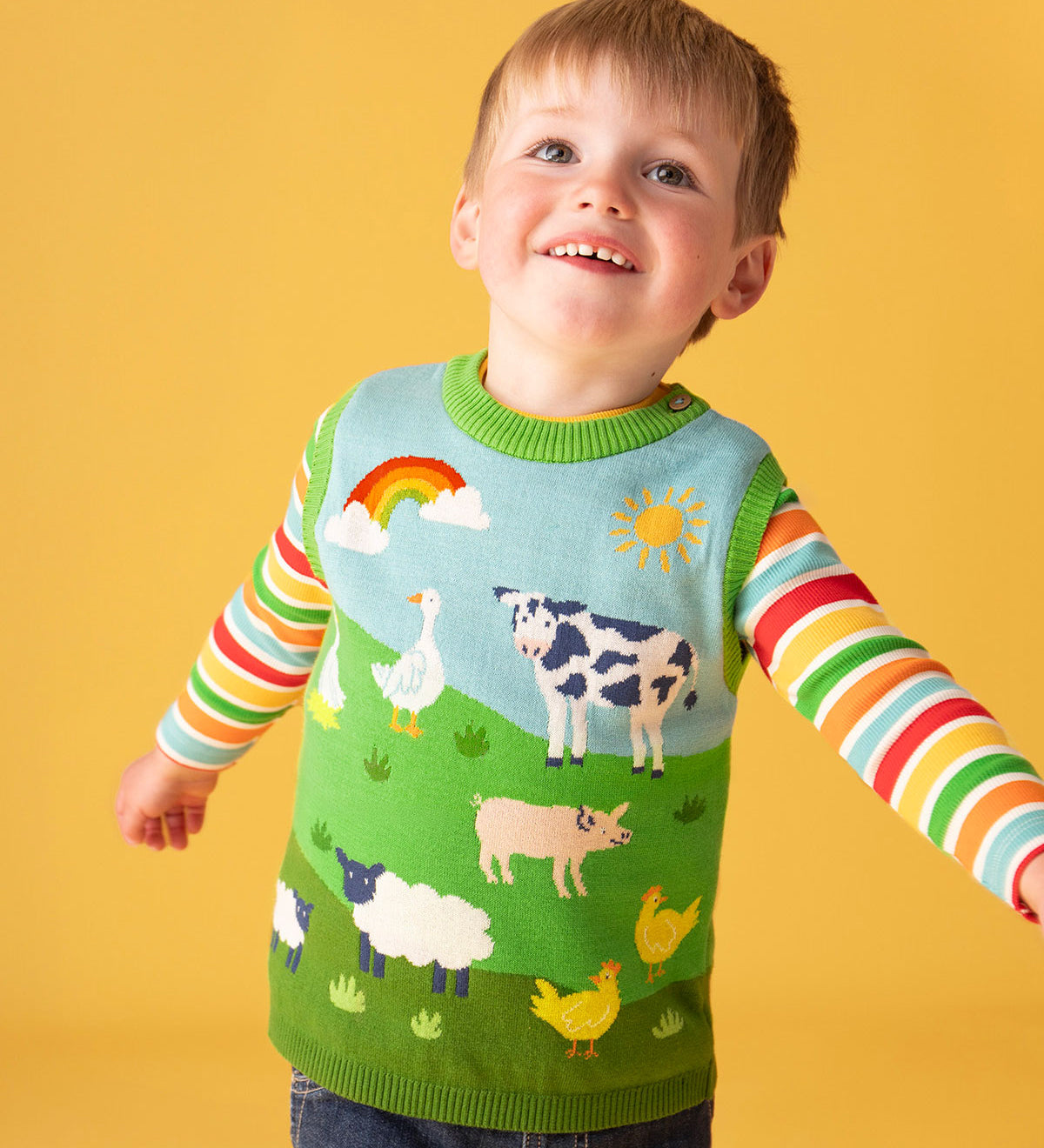A child smiling standing in front of a yellow background wearing a Frugi Organic Cotton Knitted Tank Top with a Farmyard design with a rainbow rib striped top underneath.  