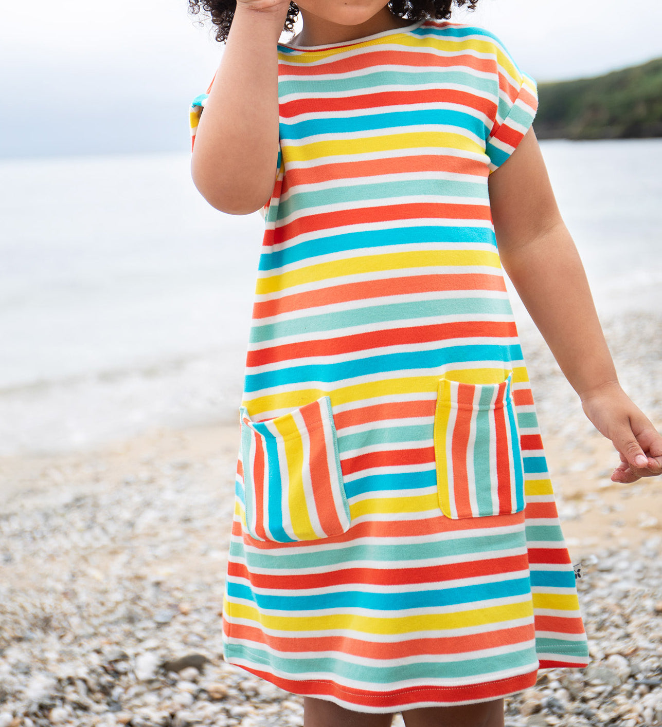 A toddler wearing the Frugi Faye Dress, featuring a Sea Rainbow Stripe print and two front pockets, standing on a pebbled beach.