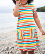 A toddler wearing the Frugi Faye Dress, featuring a Sea Rainbow Stripe print and two front pockets, standing on a pebbled beach.