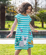 A child wearing the Frugi Felicity dress, a soft GOTS organic cotton dress with blue and white stripes, short sleeves and a grey appliqué koala on the skirt