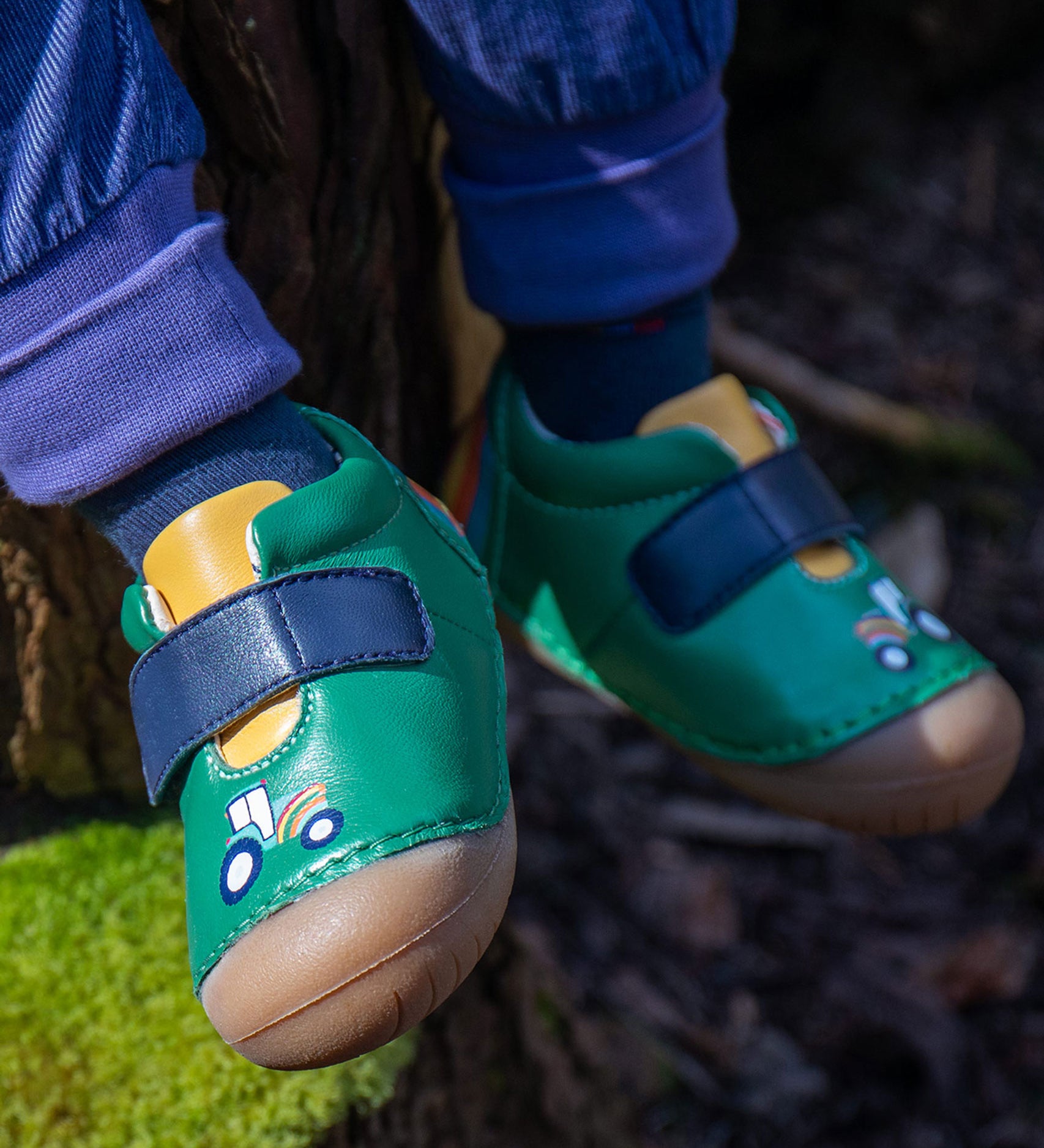 A close up of the Fjord First Walker leather baby shoes with a holly green tractor design being worn. The child is sitting outdoors.