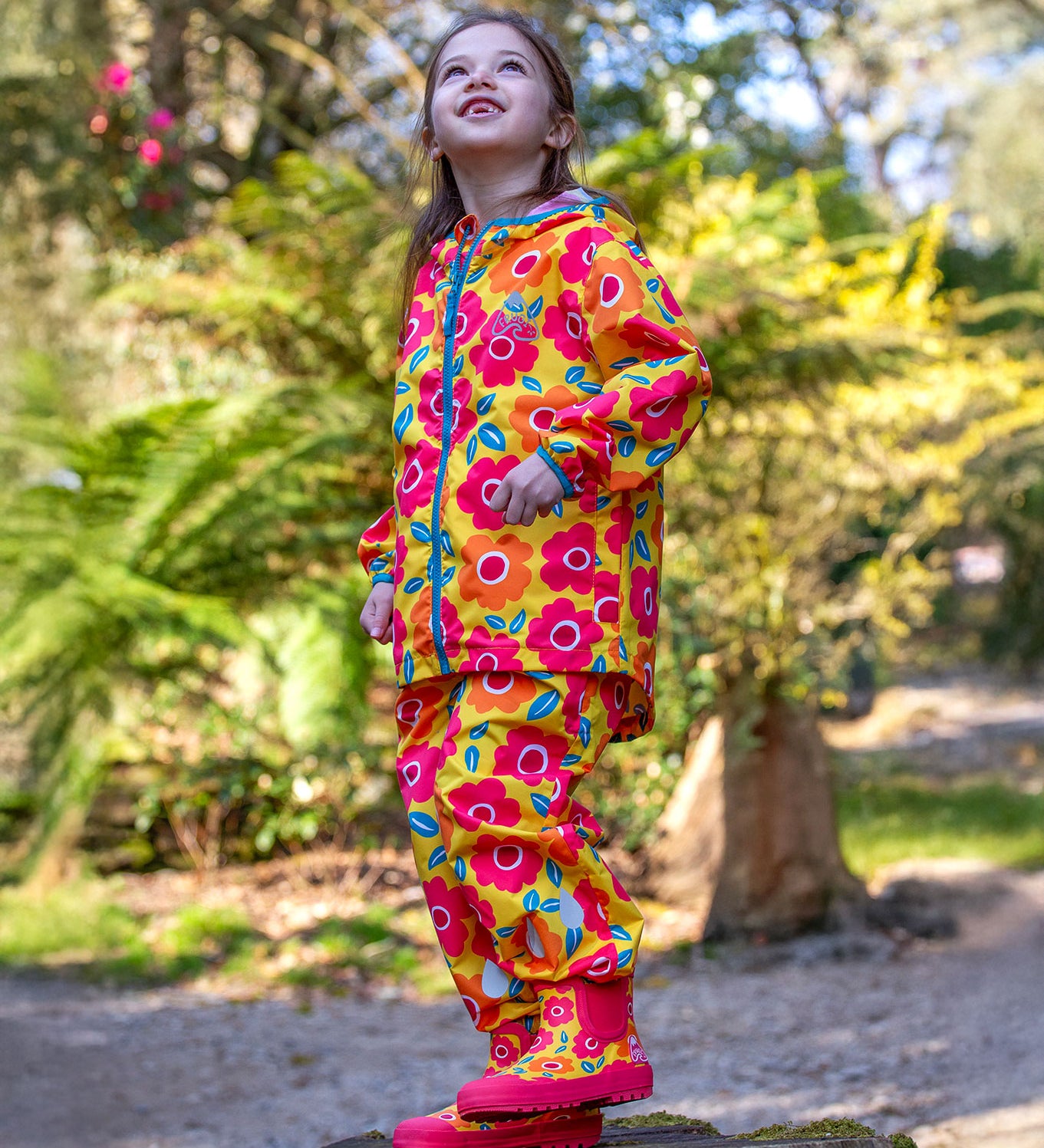 A child standing outdoors on a tree stump looking up at the sky wearing a Frugi flower fields rain jacket, waterproof trousers and matching wellington boots yellow rain or shine waterproof trousers an all over pink and orange flowers print.