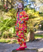 A child standing outdoors on a tree stump looking up at the sky wearing a Frugi flower fields rain jacket, waterproof trousers and matching wellington boots yellow rain or shine waterproof trousers an all over pink and orange flowers print.