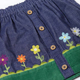 A close up of the front of the Frugi Cara cord children's skirt in ink blue with flower and grass details. The skirt has a green band with a scalloped edge to create the look of grass,