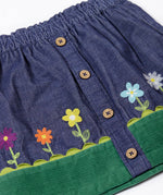 A close up of the front of the Frugi Cara cord children's skirt in ink blue with flower and grass details. The skirt has a green band with a scalloped edge to create the look of grass,